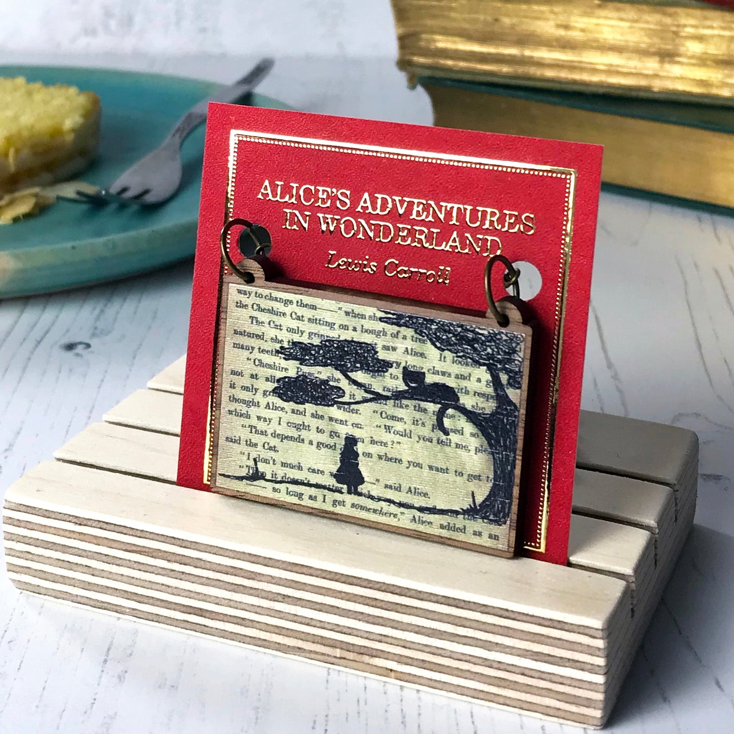 A laser cut wooden necklace by loadofolbobbins showing a silhouette scene of alice looking up at the cheshire cat in a tree against original text from the alice in wonderland novel. The necklace is mounted on a red backing card with gold foil lettering detail saying the name of the novel and writer.