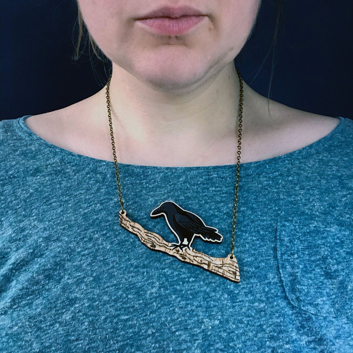 Crow necklace by Loadofolbobbins. A laser cut wooden necklace that shows an illustrated crow on a tree branch that has a pyrography textured detail. The necklace hangs on an antique bronze chain. It is being worn by me a white woman wearing a light turquoise speckled top. You can just see the bottom of my face and neck.