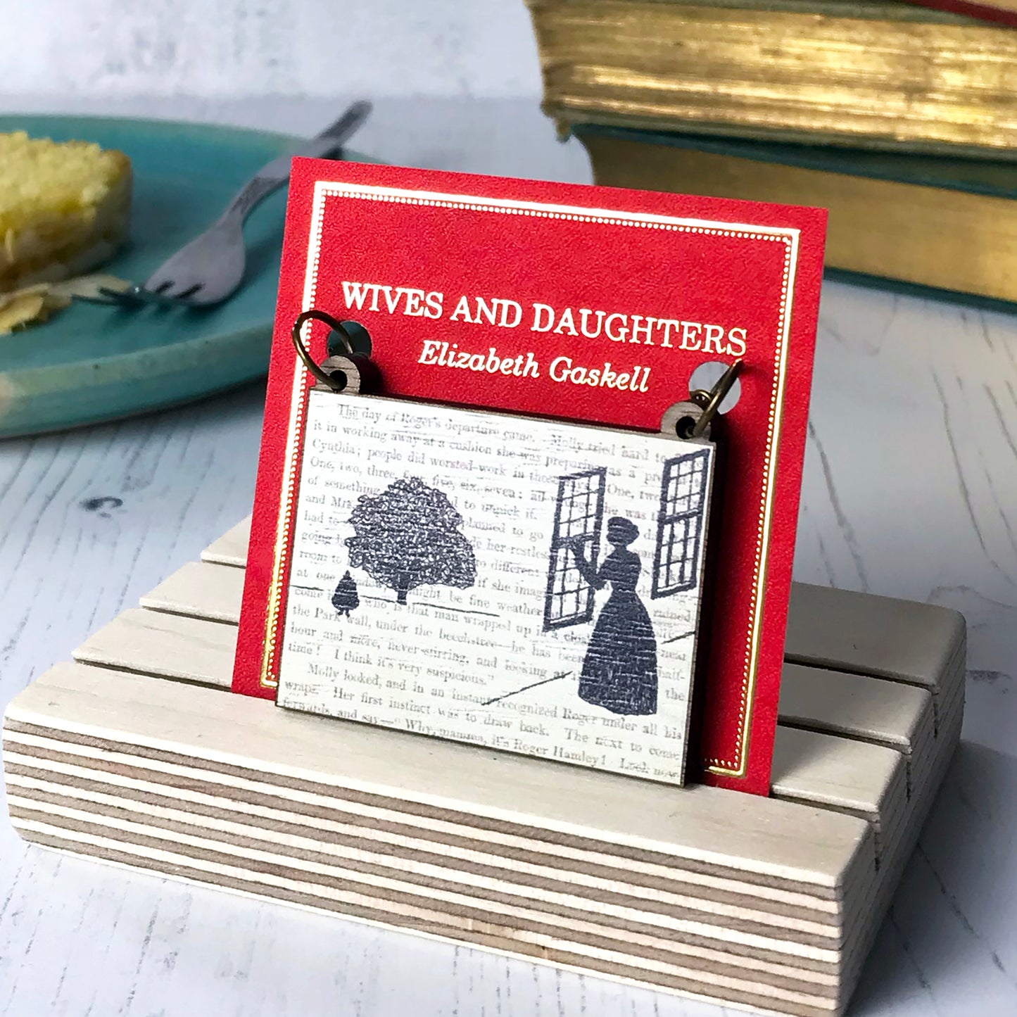 A laser cut wooden necklace by loadofolbobbins showing a silhouette scene inspired by the novel Wives and Daughters by Elizabeth Gaskell. The scene shows Molly looking out of the window at  Roger standing under a large tree. The scene is printed against original text from the novel. The necklace is mounted on a red backing card with gold foil lettering detail and made to look like the front of a book.