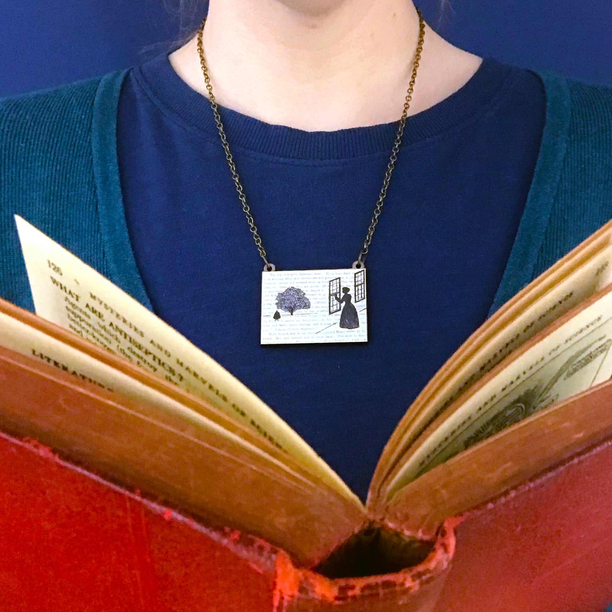 A laser cut wooden necklace by loadofolbobbins showing a silhouette scene inspired by the novel Wives and Daughters by Elizabeth Gaskell and being worn against a blue top. The scene shows Molly looking out of the window at  Roger standing under a large tree. The scene is printed against original text from the novel.